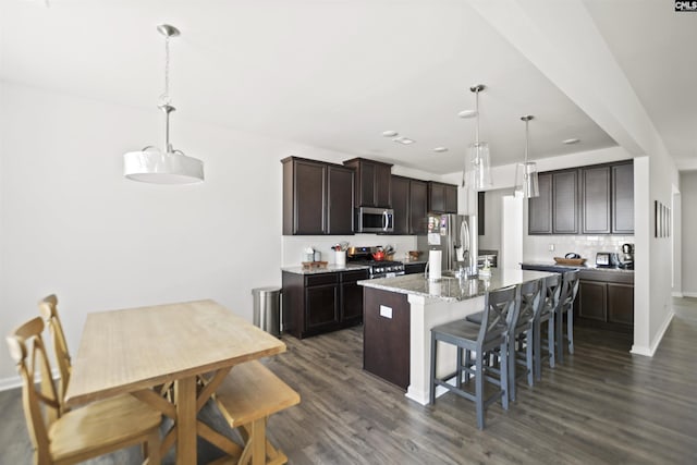 kitchen featuring appliances with stainless steel finishes, a kitchen island with sink, dark brown cabinets, hanging light fixtures, and light stone counters