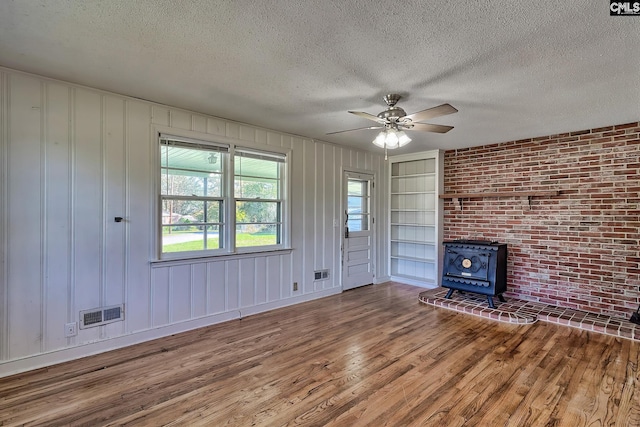 unfurnished living room with a textured ceiling, ceiling fan, wood-type flooring, built in features, and a wood stove