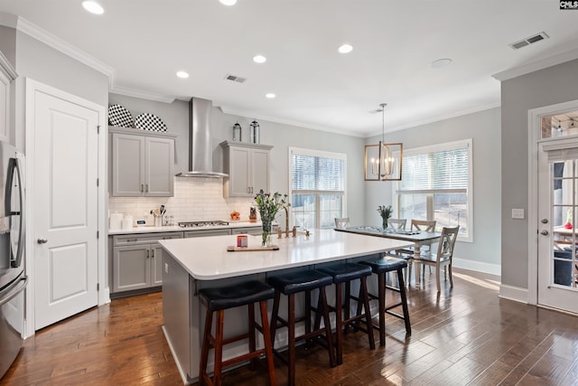 kitchen featuring gray cabinetry, a center island with sink, wall chimney exhaust hood, a breakfast bar area, and stainless steel appliances