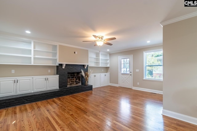 unfurnished living room featuring a brick fireplace, built in shelves, ornamental molding, ceiling fan, and wood-type flooring