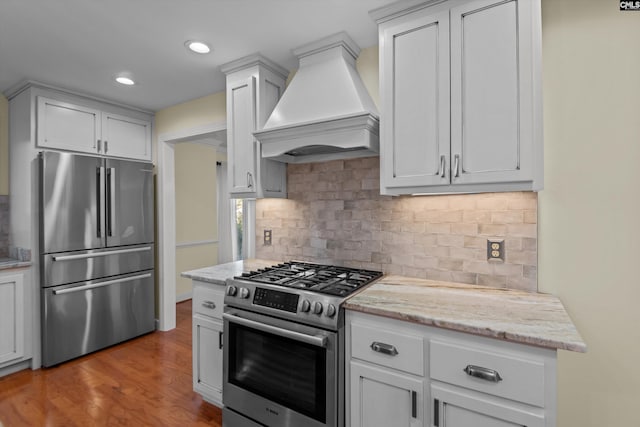 kitchen featuring light stone countertops, stainless steel appliances, decorative backsplash, light wood-type flooring, and custom range hood