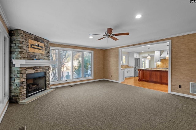 unfurnished living room featuring ceiling fan, light colored carpet, ornamental molding, and a stone fireplace