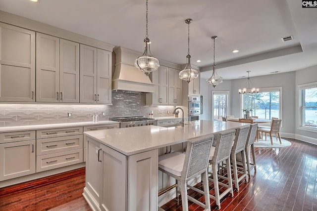 kitchen featuring premium range hood, double oven, a breakfast bar area, hanging light fixtures, and a kitchen island with sink