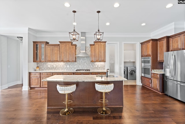 kitchen featuring appliances with stainless steel finishes, hanging light fixtures, washing machine and dryer, an island with sink, and wall chimney exhaust hood