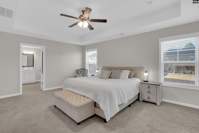 bedroom with a raised ceiling, ensuite bathroom, light colored carpet, and ceiling fan