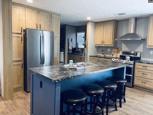 kitchen with wall chimney range hood, sink, a kitchen island with sink, stainless steel appliances, and light hardwood / wood-style floors