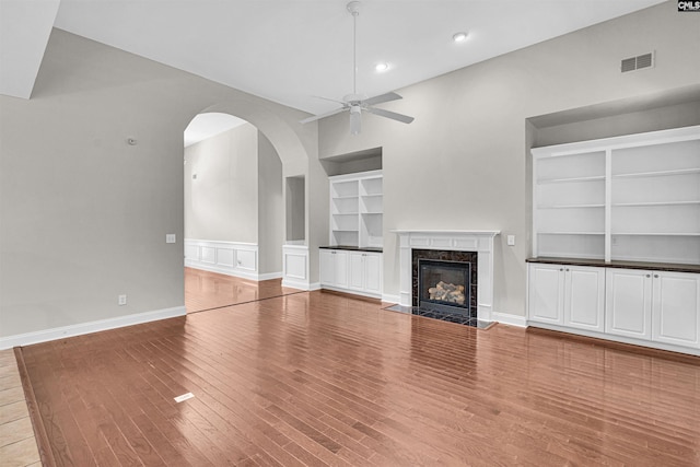 unfurnished living room with built in shelves, a high end fireplace, ceiling fan, and light wood-type flooring