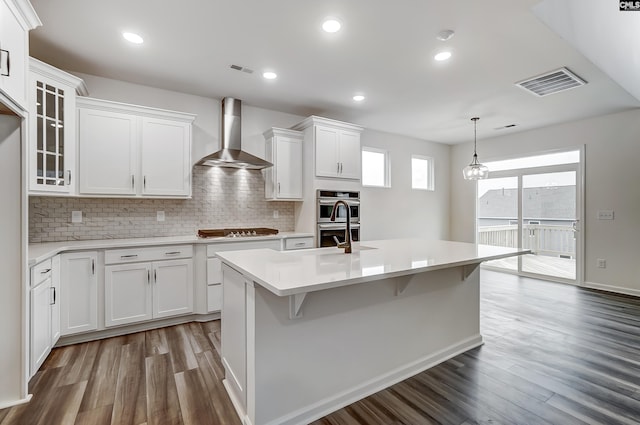 kitchen featuring wall chimney range hood, white cabinetry, stainless steel appliances, a center island with sink, and decorative light fixtures