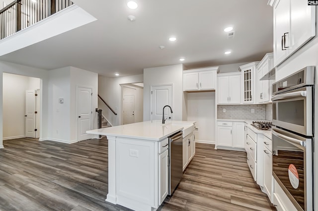 kitchen featuring hardwood / wood-style flooring, appliances with stainless steel finishes, a kitchen island with sink, backsplash, and white cabinetry