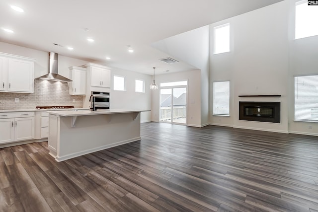 kitchen with wall chimney range hood, decorative light fixtures, white cabinets, and a center island with sink