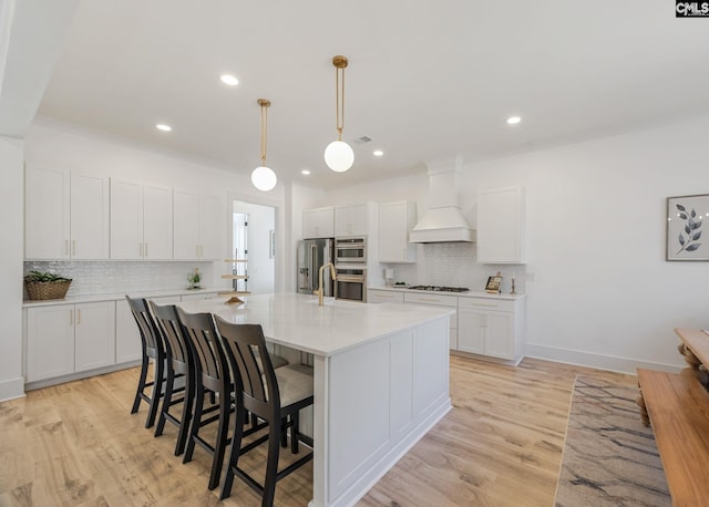 kitchen featuring premium range hood, appliances with stainless steel finishes, white cabinetry, hanging light fixtures, and a kitchen island with sink