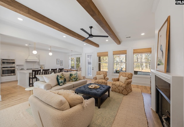 living room with ceiling fan, plenty of natural light, beamed ceiling, and light wood-type flooring