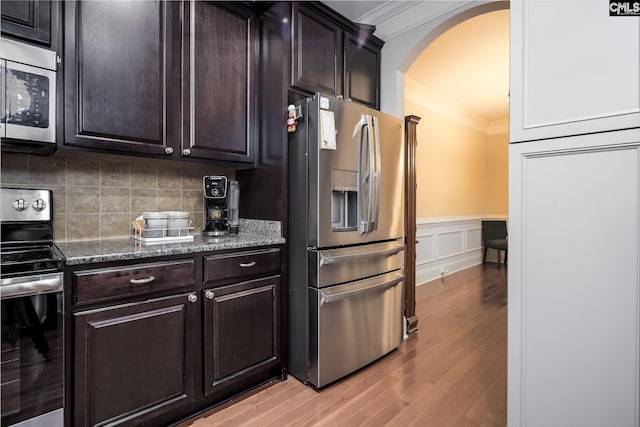 kitchen with dark brown cabinets, stainless steel appliances, ornamental molding, dark stone counters, and light wood-type flooring