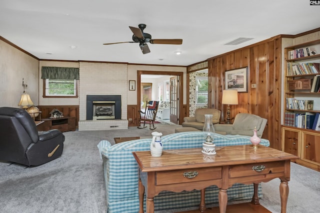 living room featuring wood walls, ceiling fan, crown molding, a brick fireplace, and light carpet