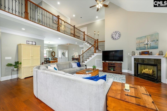 living room with ceiling fan with notable chandelier, a high ceiling, a tiled fireplace, crown molding, and dark wood-type flooring