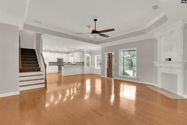 unfurnished living room with crown molding, a tray ceiling, ceiling fan with notable chandelier, and light wood-type flooring