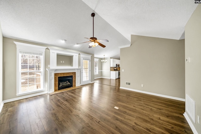 unfurnished living room with ceiling fan with notable chandelier, lofted ceiling, a tiled fireplace, dark wood-type flooring, and a textured ceiling