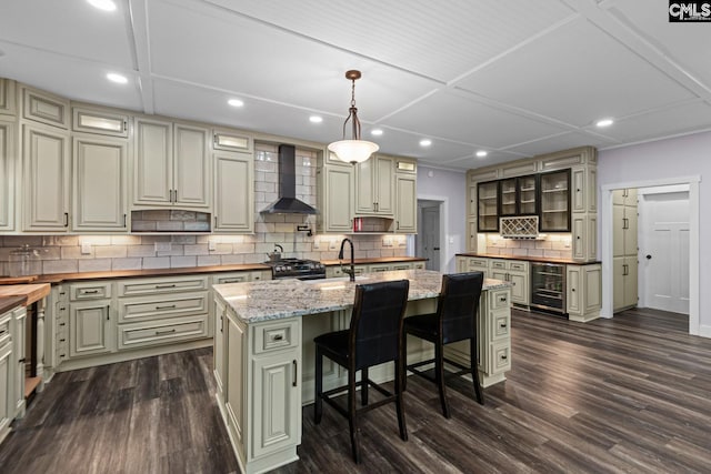 kitchen with wall chimney range hood, butcher block counters, wine cooler, an island with sink, and decorative light fixtures