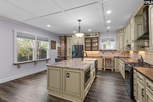 kitchen featuring gas stove, an island with sink, black fridge, decorative light fixtures, and wall chimney exhaust hood