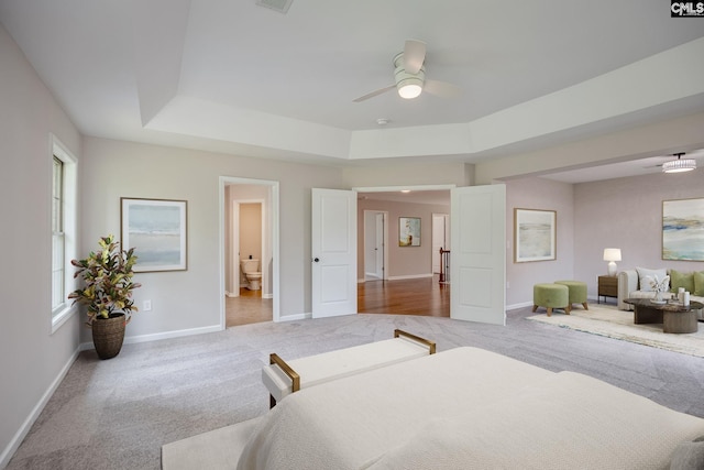 bedroom with a tray ceiling, ensuite bath, light colored carpet, and ceiling fan