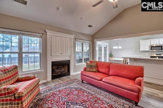 living area featuring visible vents, light wood-style floors, a large fireplace, high vaulted ceiling, and ceiling fan with notable chandelier