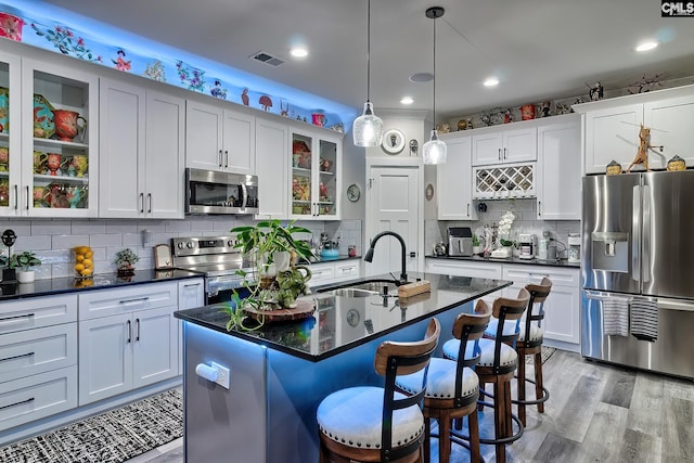 kitchen with a kitchen island with sink, appliances with stainless steel finishes, dark countertops, a kitchen bar, and glass insert cabinets