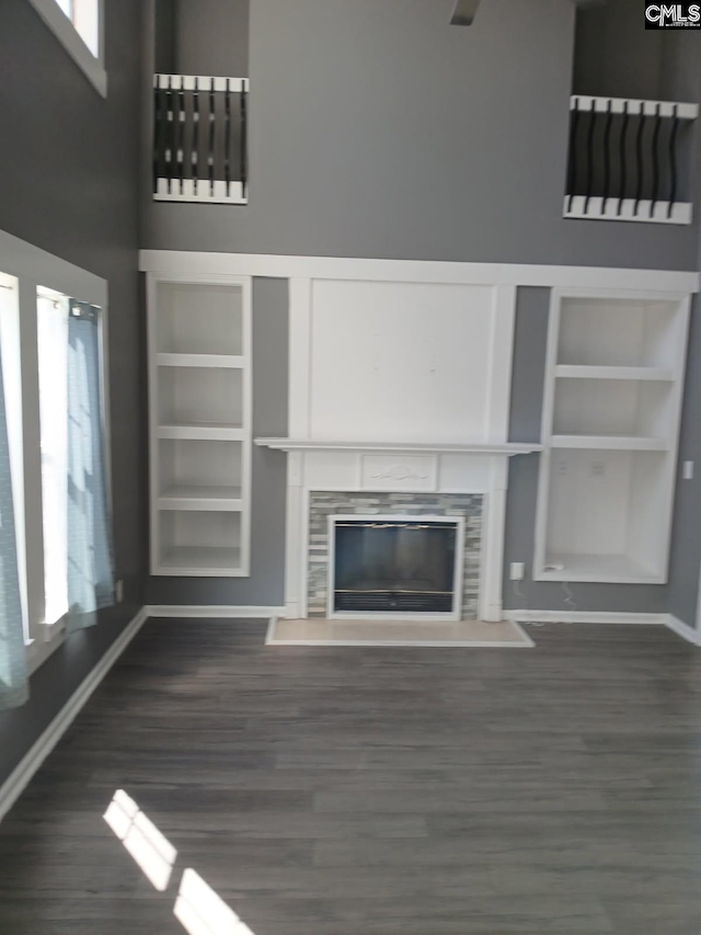 unfurnished living room with dark wood-type flooring, a towering ceiling, baseboards, built in features, and a tiled fireplace