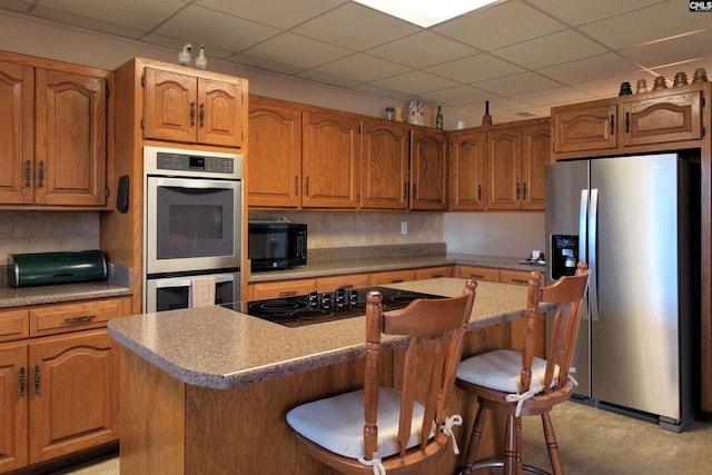 kitchen featuring brown cabinetry, a drop ceiling, a breakfast bar, a center island, and black appliances