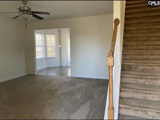 empty room featuring a ceiling fan, dark carpet, baseboards, and stairs