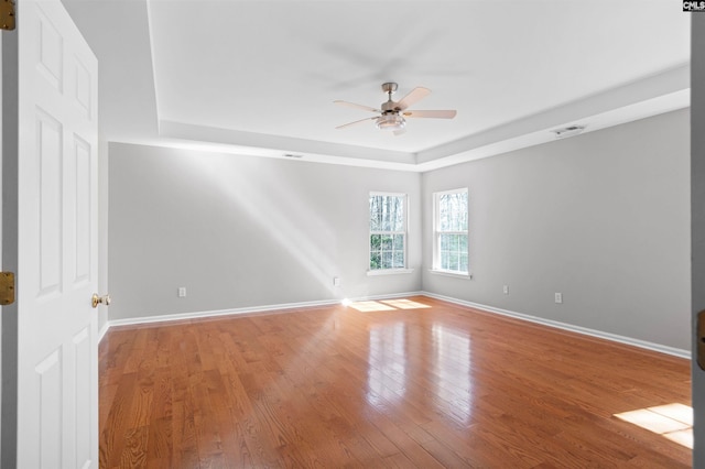 empty room with a tray ceiling, visible vents, ceiling fan, wood finished floors, and baseboards