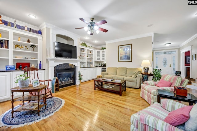 living room featuring built in features, a ceiling fan, a glass covered fireplace, light wood-style flooring, and crown molding