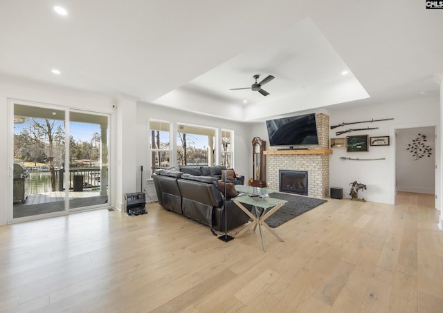 living room featuring a brick fireplace, a raised ceiling, ceiling fan, a water view, and light wood-style floors