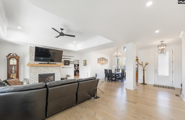 living room with a brick fireplace, a raised ceiling, light wood finished floors, and ceiling fan with notable chandelier