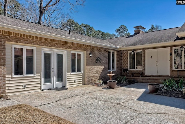 view of exterior entry featuring brick siding, a shingled roof, french doors, a chimney, and a patio area