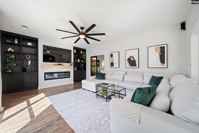 living room with ceiling fan, built in shelves, wood finished floors, visible vents, and a glass covered fireplace