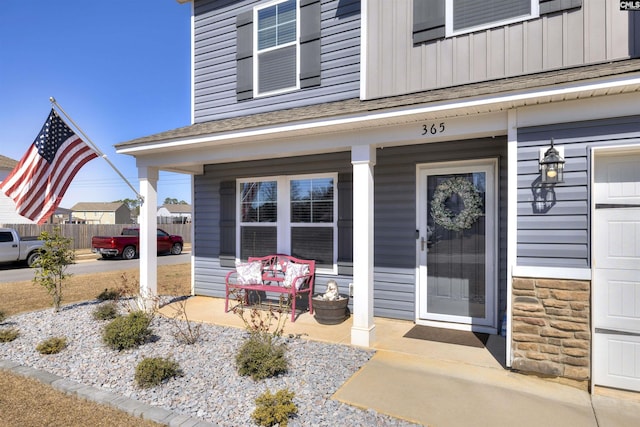 view of exterior entry featuring a shingled roof, covered porch, board and batten siding, fence, and stone siding