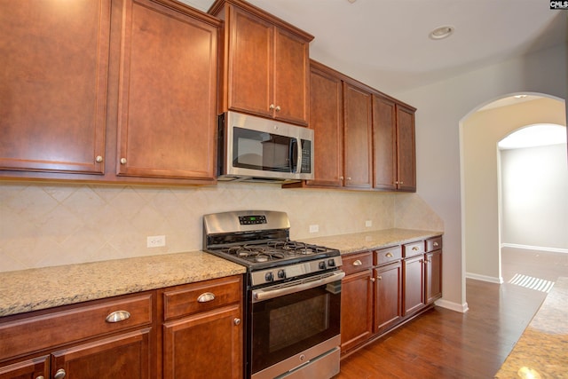 kitchen featuring arched walkways, light stone counters, stainless steel appliances, baseboards, and backsplash