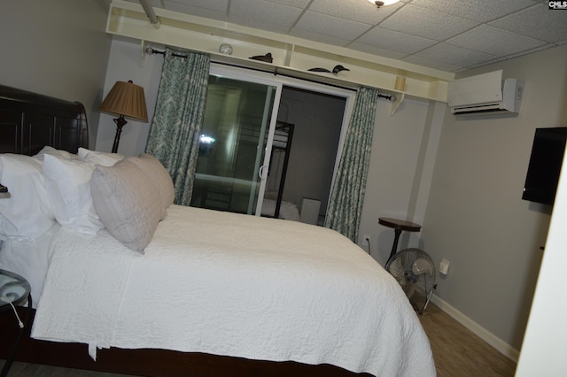 bedroom featuring baseboards, a wall mounted air conditioner, a drop ceiling, and wood finished floors