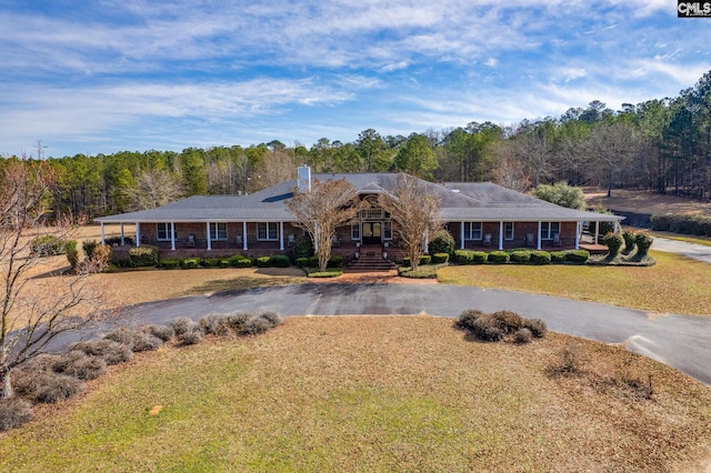 ranch-style house with brick siding, a chimney, a view of trees, driveway, and a front lawn