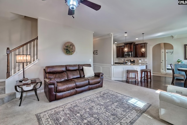 carpeted living room with stairway, arched walkways, a decorative wall, and a ceiling fan