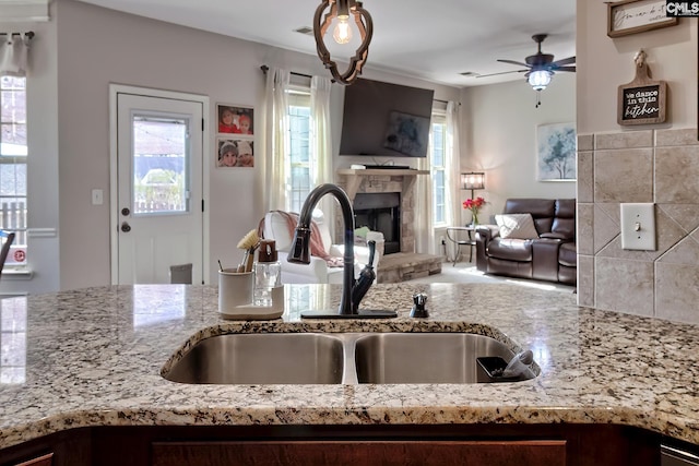 kitchen with a fireplace with raised hearth, a ceiling fan, light stone counters, open floor plan, and a sink