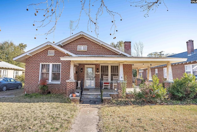 bungalow-style home with covered porch, brick siding, a front lawn, and a chimney