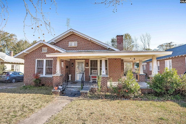 bungalow featuring covered porch, brick siding, a chimney, and a front yard