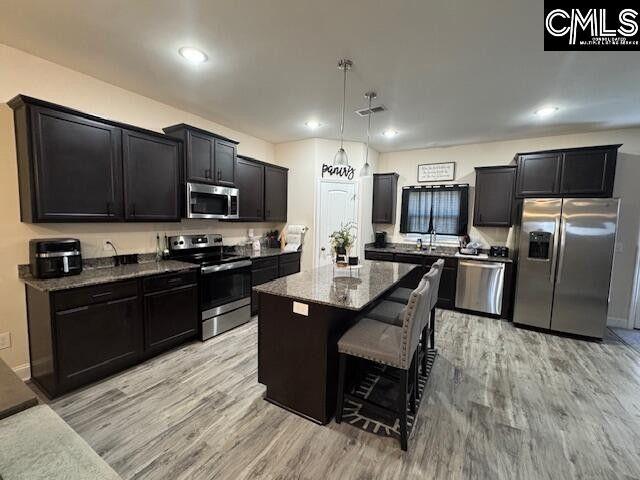 kitchen featuring appliances with stainless steel finishes, dark cabinets, dark stone countertops, a center island, and light wood-type flooring