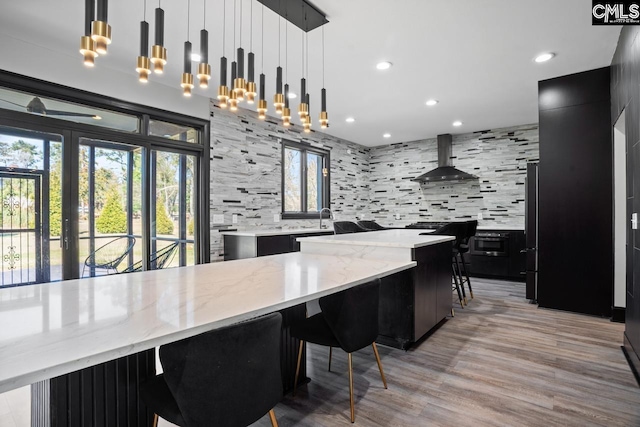 kitchen with tasteful backsplash, wall chimney exhaust hood, a breakfast bar area, dark cabinets, and light wood-type flooring