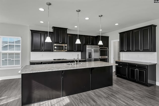 kitchen featuring stainless steel appliances, a sink, dark cabinetry, backsplash, and a large island with sink