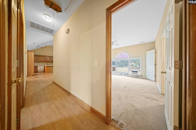 hall featuring attic access, visible vents, baseboards, light wood-style flooring, and vaulted ceiling