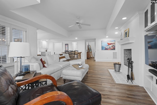 living room featuring a fireplace with flush hearth, a tray ceiling, recessed lighting, wood finished floors, and a ceiling fan