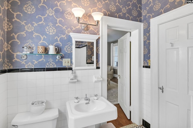 half bath with toilet, a sink, tile walls, wainscoting, and wallpapered walls