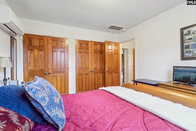 bedroom with two closets, visible vents, a textured ceiling, and a wall mounted air conditioner